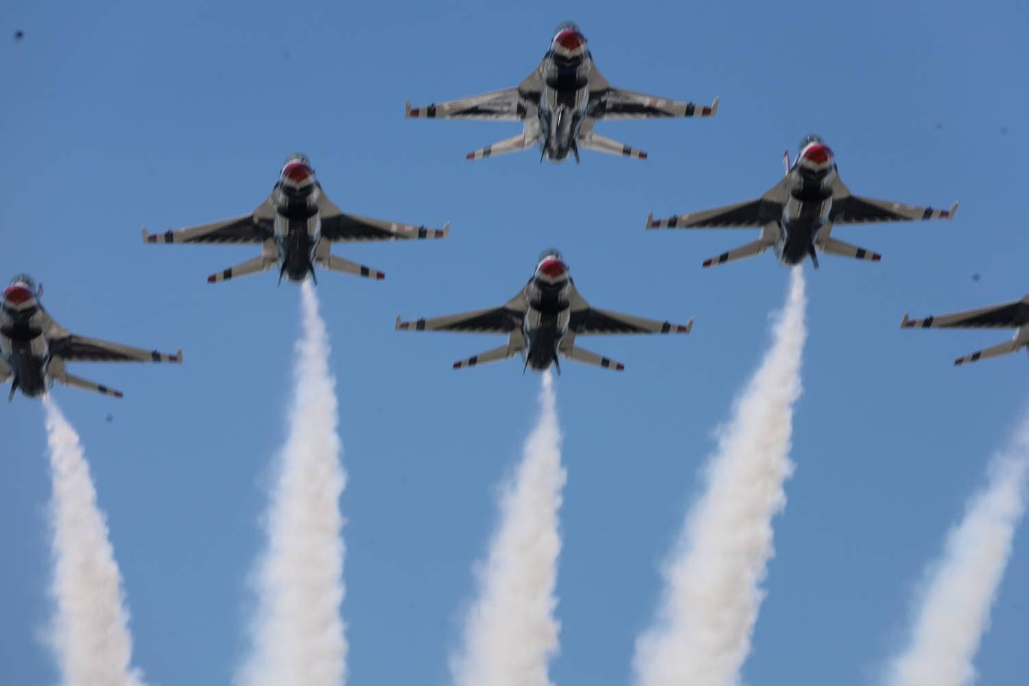 Five fighter jets in a synchronized formation releasing white smoke trails into a clear blue sky.