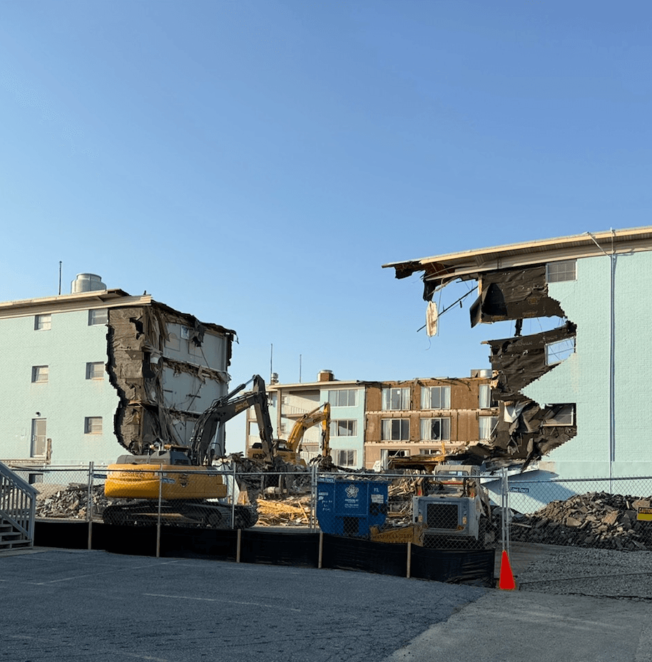 Construction site: a yellow excavator demolishes a pale-blue apartment building, with collapsed walls and rubble behind a chain-link fence under a clear blue sky.