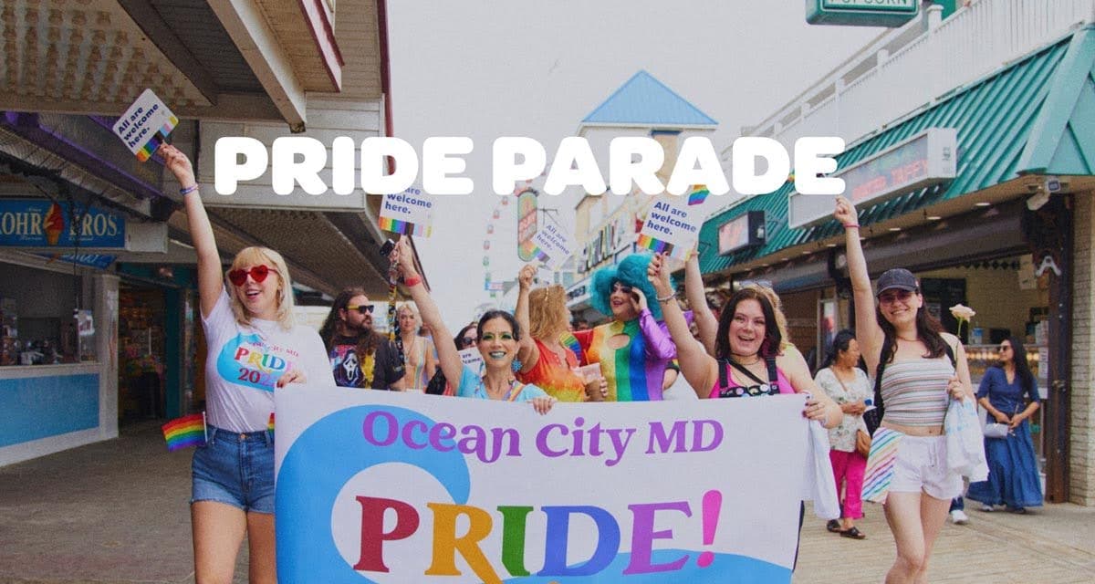 Parade participants hold a banner reading Ocean City MD PRIDE as they march down a street with rainbow signs.